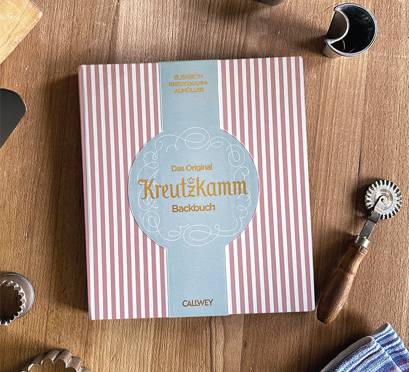Buch liegt auf einem Holztisch, umgeben von Backutensilien. Text: "Das Original Kreutzkamm Backbuch"