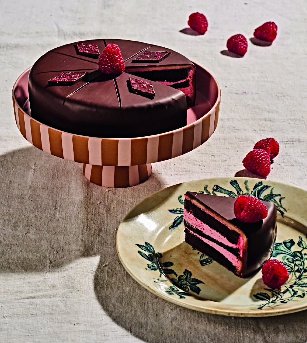 Eine Schokoladentorte mit Himbeeren steht auf einem Kuchenstaender, umgeben von mehreren Himbeeren auf dem Tisch. Ein Stueck der Torte liegt auf einem Teller daneben.